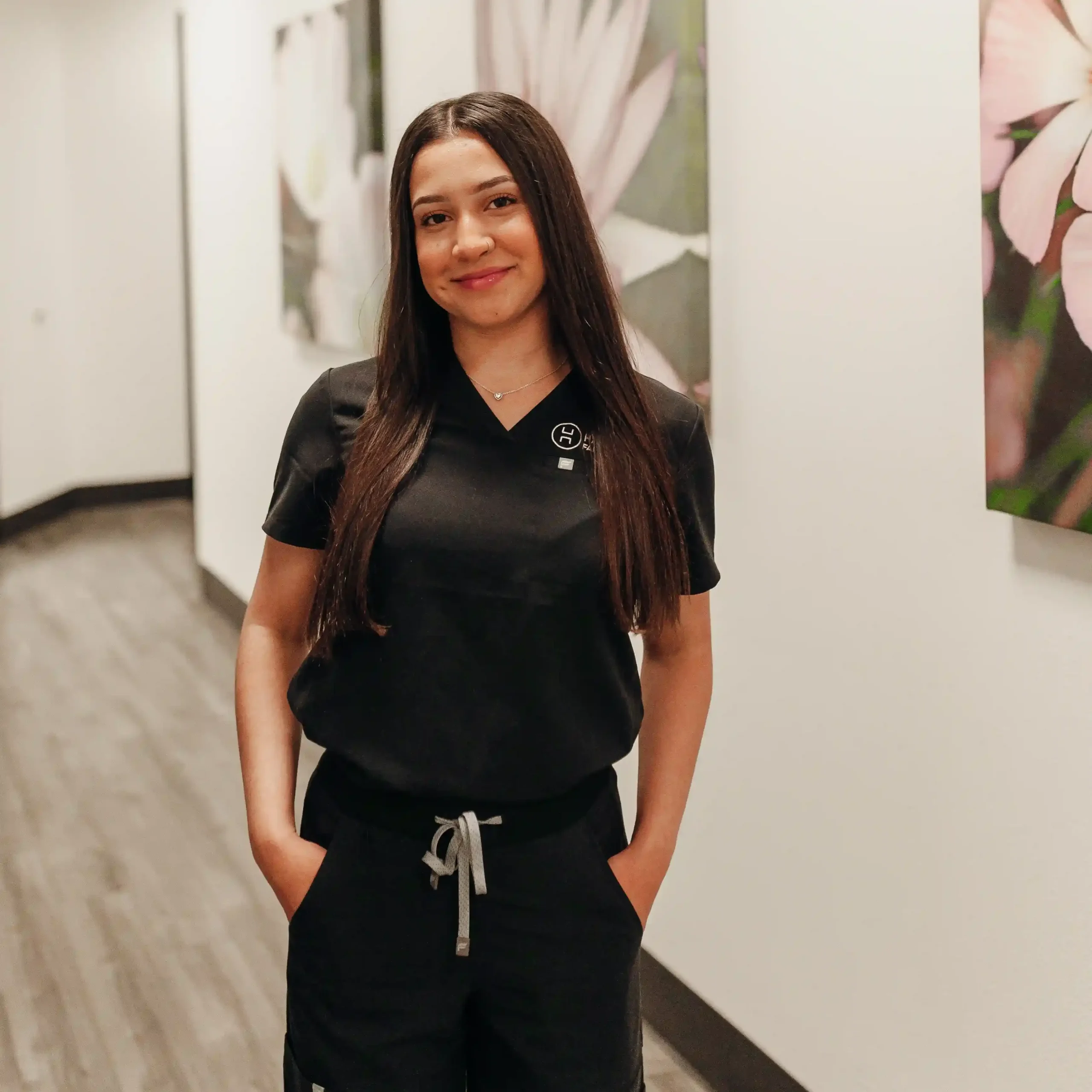 Clinical care team member Jasmin Pantoja smiling in black medical scrubs inside a well-lit clinic office corridor.